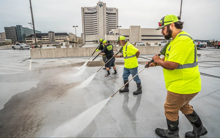 Four workers in PPM uniform and caps pressure wash a concrete parking lot